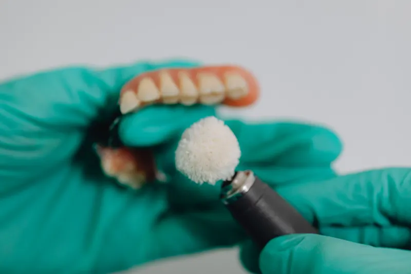 Gloved hands polish a set of false teeth with a small rotating brush.