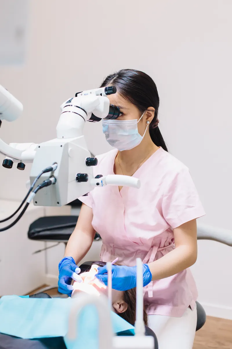 A dentist in pink scrubs looks through a dental microscope beside the treatment chair.